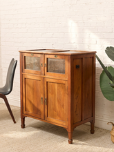 Load image into Gallery viewer, A mid-century modern wooden cabinet with a warm medium-brown finish, featuring two upper glass-front drawers and two lower doors, styled in a room against a white brick wall.
