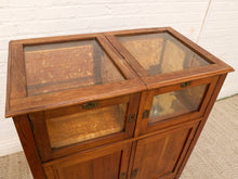 Load image into Gallery viewer, A vintage wooden display cabinet with glass doors and a herringbone wood top, photographed against a white brick wall and light-colored carpet.
