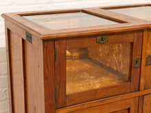 Load image into Gallery viewer, A close-up of a vintage wooden display cabinet with glass doors and a glass-topped surface, set against a white brick wall.

