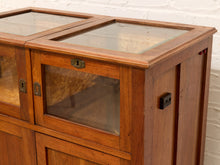 Load image into Gallery viewer, A close-up of a wooden display cabinet with glass doors and drawers, showing warm medium-brown wood grain and brass hardware against a white brick wall background.
