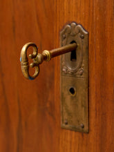 Load image into Gallery viewer, An antique brass key with a decorative loop handle protruding from an old bronze lockset on a warm brown wooden door.
