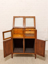 Load image into Gallery viewer, A mid-century wooden cabinet with glass-front upper doors, open drawers, and open lower doors, set against a white brick wall and light carpet.
