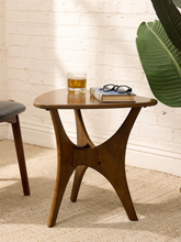 Load image into Gallery viewer, A small wooden side table with a rounded top and sculptural curved legs, styled in a room setting with a glass of beer, a book, and eyeglasses on top, set against a white brick wall and a large green leaf.
