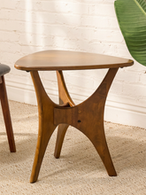 Load image into Gallery viewer, A mid-century modern wooden side table with a triangular top and sculptural curved legs, shown in a styled room setting with a white brick wall and woven rug.
