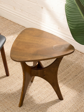 Load image into Gallery viewer, A mid-century modern triangular wooden side table with a warm brown finish, shown on a textured beige rug next to a white brick wall and a green plant leaf.
