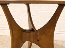 Load image into Gallery viewer, Close-up of a mid-century modern wooden table base with a warm brown finish, featuring a distinctive curved T-leg design and a glass top edge, set against a white brick wall and light carpet.
