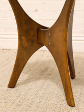 Load image into Gallery viewer, Close-up of a wooden table base with four splayed legs and a central joint, shown on a textured beige rug against a white brick wall.
