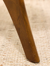 Load image into Gallery viewer, Close-up of a wooden chair leg with a warm brown finish resting on a textured beige woven rug.
