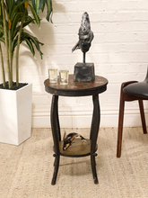 Load image into Gallery viewer, A small round wooden end table with a dark metal frame and lower shelf, styled with a bird sculpture, two small glasses, and a decorative object. The table is set on a beige rug against a white brick wall next to a potted plant and a chair.
