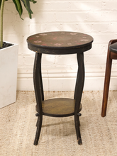 Load image into Gallery viewer, A small round dark wood side table with a distressed finish, featuring a top shelf and a floral pattern on the tabletop, set against a white brick wall.
