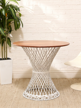 Load image into Gallery viewer, A round side table with a warm brown wood top and a white woven wire pedestal base, styled in a bright room with a white brick wall, a potted plant, and a partial cream chair.
