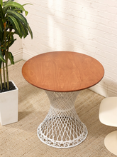 Load image into Gallery viewer, A round wooden end table with a white woven wire pedestal base, styled in a bright room with a white brick wall, beige rug, and a potted plant.
