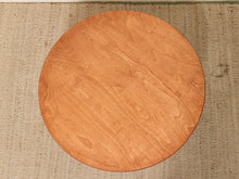 Load image into Gallery viewer, A round wooden board or tray with a warm honey-brown wood grain finish, displayed flat on a textured beige woven rug.
