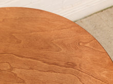 Load image into Gallery viewer, Close-up of a round wooden table showing a warm medium-brown wood grain finish, with a white baseboard and light rug in the background.
