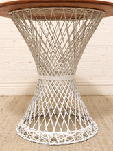 Load image into Gallery viewer, A mid-century modern wire chair or side table base with a white woven metal structure and a round wooden top, set against a white brick wall and beige rug.
