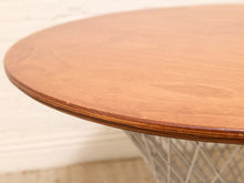 Load image into Gallery viewer, Close-up of a round wooden table top with a warm medium-brown finish and visible wood grain, showing part of the metal wire base in a real interior setting.
