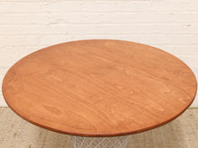 Load image into Gallery viewer, A round wooden coffee table with a warm medium-brown cherry wood finish and a white wire pedestal base, set against a white brick wall.
