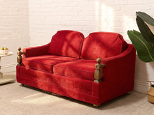 Load image into Gallery viewer, A red velvet loveseat sofa with turned wooden armrests, positioned in a room with a white brick wall, beige carpet, and a green potted plant.
