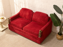 Load image into Gallery viewer, A red velvet loveseat sofa with wooden armrests, positioned in a room corner against a white brick wall with a potted plant and a side table nearby.
