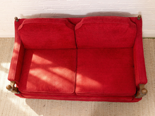 Load image into Gallery viewer, A red upholstered loveseat or sofa viewed from above, with two seat cushions and two back cushions, set against a white brick wall and light beige rug.
