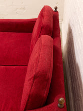 Load image into Gallery viewer, A close-up of a red upholstered armchair or sofa corner with plush cushions and small round wooden finials on the arm ends, set against a white brick wall.
