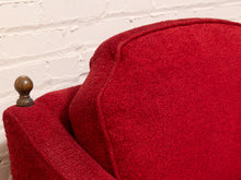 Load image into Gallery viewer, Close-up of a red upholstered armchair corner with a dark wooden finial on the arm, set against a white painted brick wall.
