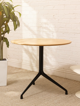 Load image into Gallery viewer, A round cafe table with a light natural wood top and a black metal pedestal base with three legs, set against a white brick wall and beige carpet next to a potted plant.

