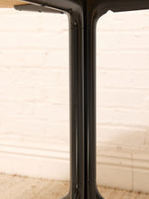 Load image into Gallery viewer, Close-up of a glossy black metal table pedestal base with a light wood tabletop edge, set against a white painted brick wall and beige carpet.
