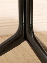 Load image into Gallery viewer, Close-up of a glossy black metal tripod base with three legs converging at a central joint, set against a softly blurred light beige wall and carpet.
