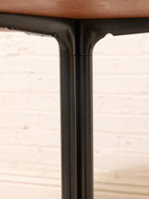 Load image into Gallery viewer, Close-up of a black metal table leg joint supporting a warm brown wood tabletop, with a softly blurred white brick wall background.
