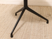Load image into Gallery viewer, Close-up of a black metal table base with three legs, resting on a beige woven rug against a white wall.
