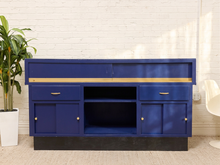 Load image into Gallery viewer, A blue wooden console cabinet with gold handles and knobs, set against a white brick wall with a potted plant on the left and a white chair on the right.
