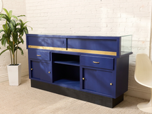 Load image into Gallery viewer, A royal blue wooden sideboard cabinet with gold trim and brass hardware, featuring a glass-top aquarium display case on the right, set against a white brick wall with a potted plant and a white chair.
