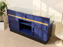 Load image into Gallery viewer, A blue display case or cabinet with a clear glass top, gold trim, and brass-tone hardware, shown in a styled room setting with a white brick wall, beige carpet, a potted plant, and a white chair.
