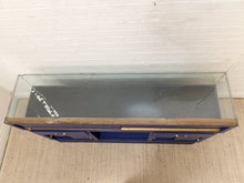 Load image into Gallery viewer, A blue wooden display case or cabinet with a glass top and brass handles, photographed from above against a white brick wall and beige carpet.
