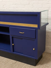 Load image into Gallery viewer, A blue wooden media console or aquarium stand with a glass tank on top, featuring a gold stripe, a drawer with a gold handle, and cabinet doors with gold knobs, set against a white brick wall.
