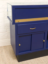 Load image into Gallery viewer, A blue wooden cabinet with a glass top, gold trim, and gold hardware, shown in a room setting against a white brick wall and beige carpet.
