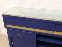 Load image into Gallery viewer, A close-up of a blue wooden display case or cabinet with a clear glass top, brass-toned hardware, and a gold horizontal trim stripe, set against a white brick wall.
