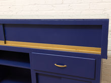 Load image into Gallery viewer, A close-up of a blue wooden cabinet or desk with a gold drawer pull and a gold horizontal trim line, set against a white brick wall background.
