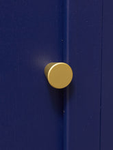 Load image into Gallery viewer, A round, matte gold metal cabinet knob mounted on a dark blue wooden door.
