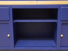 Load image into Gallery viewer, A close-up of a blue wooden cabinet or sideboard with an open center compartment, a single shelf, and small round gold knobs on the side panels.
