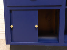 Load image into Gallery viewer, A close-up of a blue cabinet or shelf unit with a sliding door and two round gold knobs, set against a light wall and carpeted floor.
