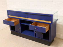 Load image into Gallery viewer, A blue wooden display cabinet with a glass top, two open drawers showing natural wood interiors, and open shelving below, photographed against a white brick wall and light carpet.
