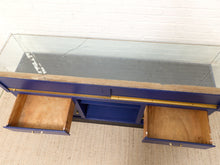 Load image into Gallery viewer, A vintage blue wooden display cabinet with a clear glass top, featuring two open drawers on the front with natural wood interiors and brass handles, set against a white brick wall and light-colored carpet.
