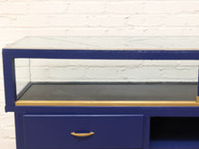 Load image into Gallery viewer, A blue display case or cabinet with a glass top, a black interior shelf, and brass-colored handles, set against a white brick wall.
