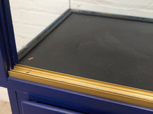 Load image into Gallery viewer, Close-up of a blue cabinet or desk frame with a black tabletop and a gold metal trim strip along the edge, set against a white brick wall background.
