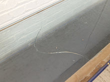 Load image into Gallery viewer, A close-up of a large clear glass panel with a visible crack running diagonally across the surface, set against a white brick wall and a tan floor or baseboard.

