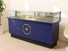 Load image into Gallery viewer, A vintage-style blue display case with a glass top case, featuring a gold octagonal emblem on the front and a black base, set against a white brick wall.
