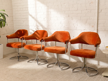 Load image into Gallery viewer, Four mid-century modern swivel chairs with orange velvet upholstery and chrome bases, arranged in a row against a white brick wall with sunlight casting shadows.
