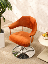 Load image into Gallery viewer, An orange velvet swivel dining chair with a chrome metal base, set in a bright room with a white brick wall, a potted plant, and a small side table.
